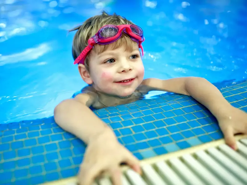 young child holding onto the side of a swimming pool with goggles on head