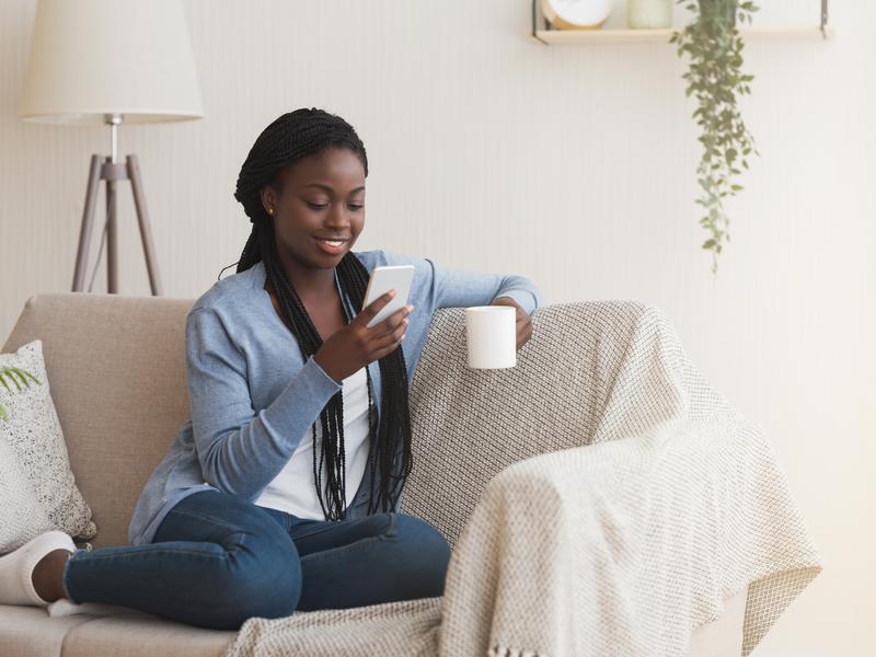 Young woman sitting on a couch, holding a coffee mug in one hand while messaging her provider on her mobile phone with the other hand