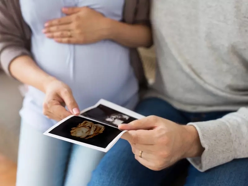  Pregnant woman sitting next to her partner, placing a hand on her belly while they both look at an ultrasound image of their baby.