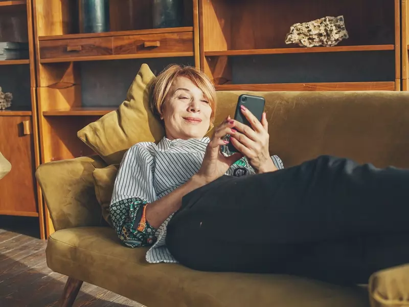 Woman lying on a couch, smiling at her mobile phone while scheduling a telehealth visit online