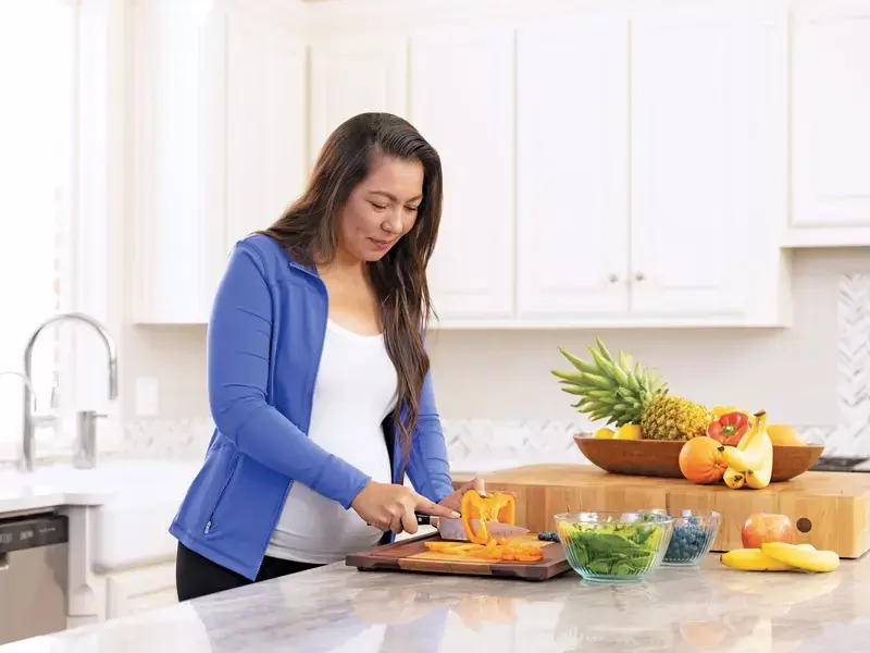 Pregnant woman standing in the kitchen, carefully cutting vegetables as part of her healthy maternity care lifestyle.