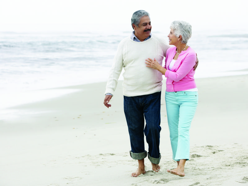 Woman living with urogynecology issues smiling and embracing her husband while they walk together on a cool day at the beach