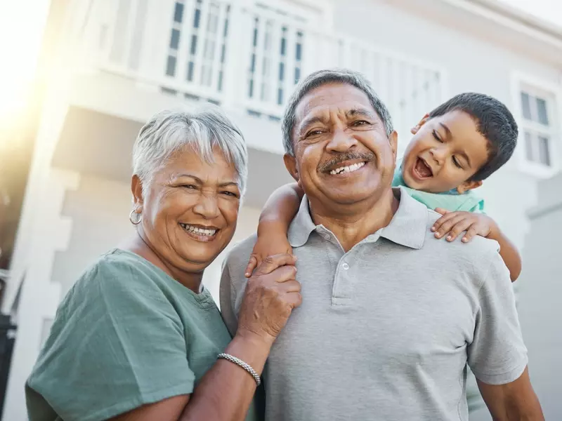 Two grandparents smile outdoors while posing with their grandchild on his grandfather's shoulders