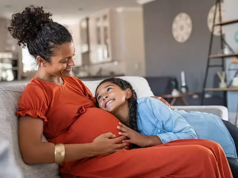 Expectant mother seated on the couch, sharing a smile with her young daughter in their living room