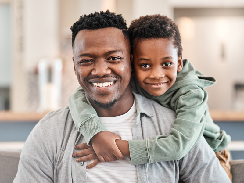 A young boy wraps his hands around his dads shoulder as they pose for a photo
