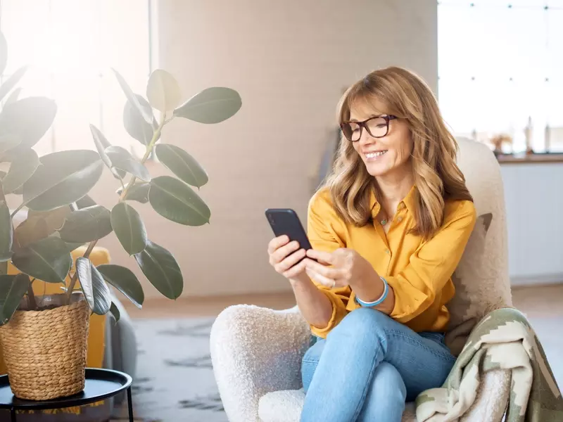 Woman seated indoors in her living room, smiling as she looks at her mobile phone while completing a virtual care questionnaire