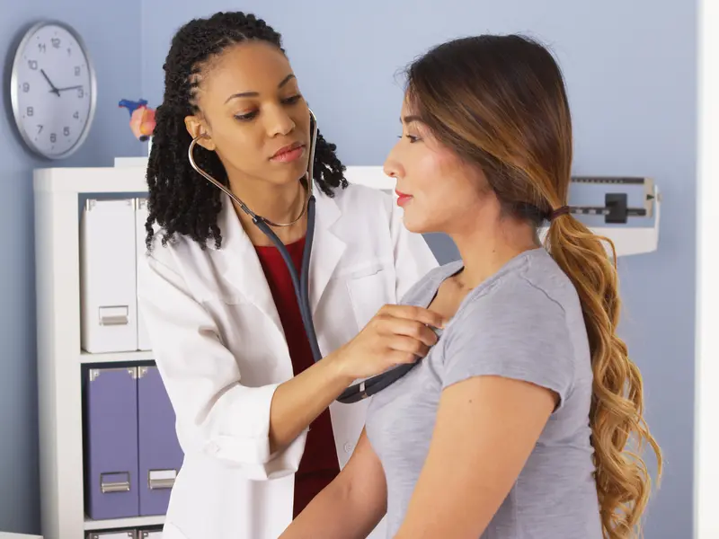 Female doctor using stethoscope to examine female patient's heart for symptoms of angina