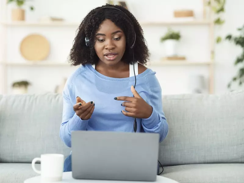 Young woman sitting on a couch wearing headphones, engaged in a conversation with her virtual care provider on her laptop