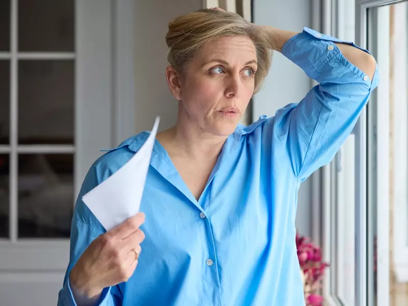 Woman standing in front of an open window in her home, fanning herself with her hand as she experiences hot flashes during the menopause transition