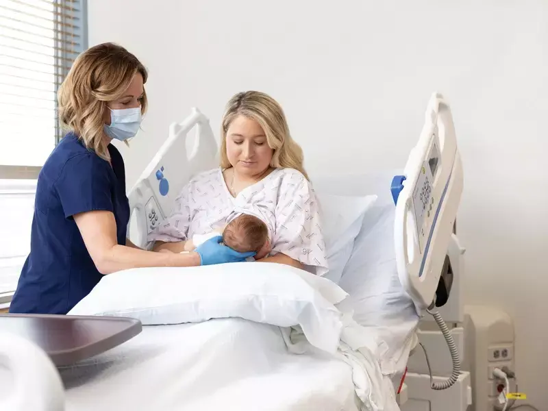 Lactation consultant assisting a new mother with breastfeeding her newborn infant while seated in a labor and delivery hospital bed