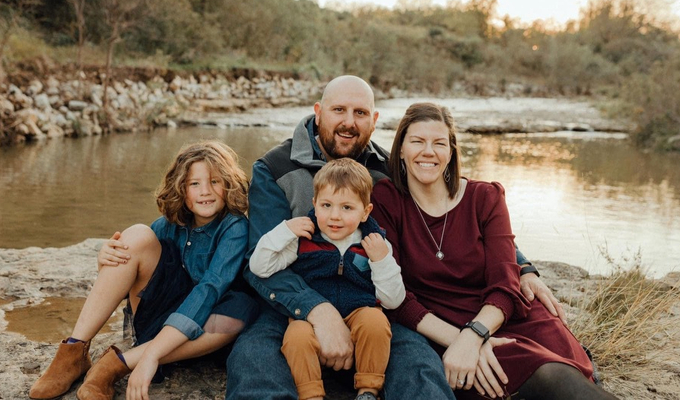 Katie Blalock poses for a family photo with her husband and two children outside near a river