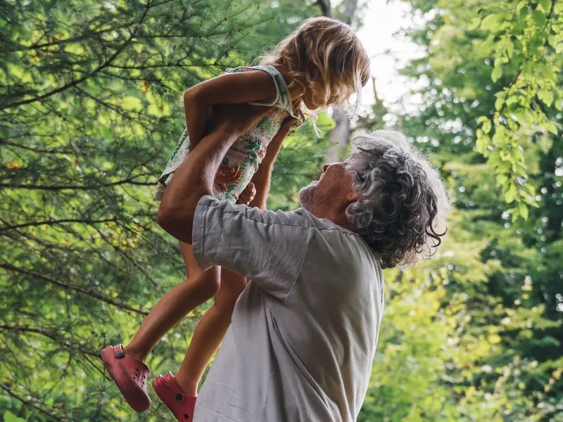 older man holding up young child in the air outside by trees on sunny day