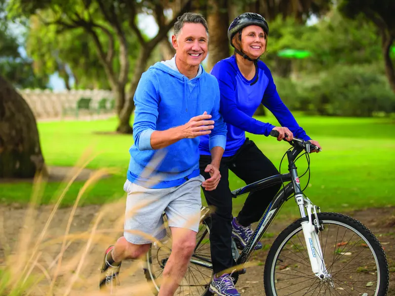 Woman going through menopause riding a bike with her husband jogging alongside her on a cool, sunny day