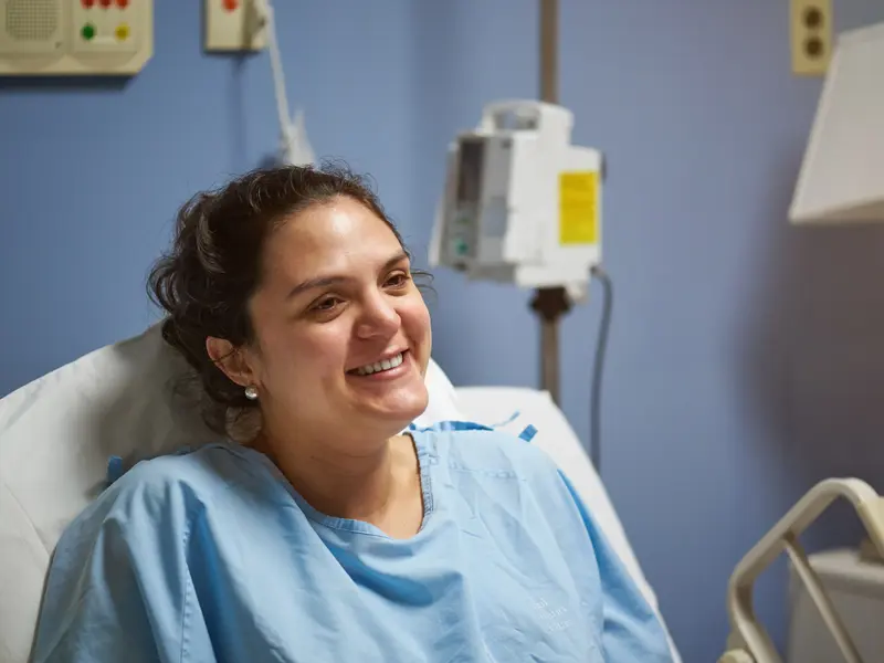 Woman in a hospital bed wearing a hospital gown, smiling off-camera while preparing for sternotomy surgery