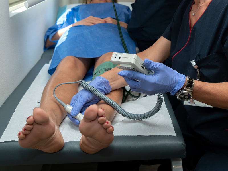  Woman in a medical gown lying on an exam table as a healthcare provider performs an ankle-brachial index on her legs