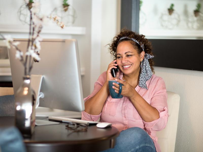 Woman seated at a desk, holding a mug and smiling while talking on her mobile phone with a virtual headache care provider