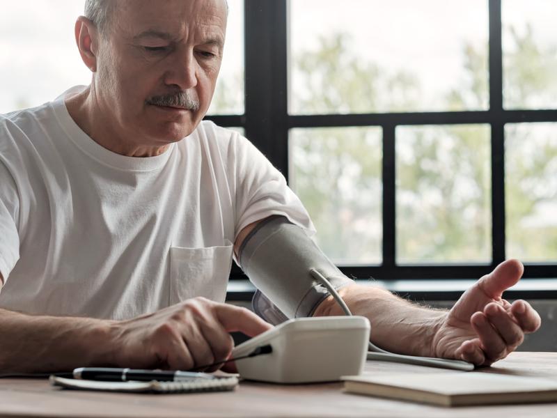 Elderly man seated at a table using a blood pressure monitor to monitor high blood pressure associated with congestive heart failure