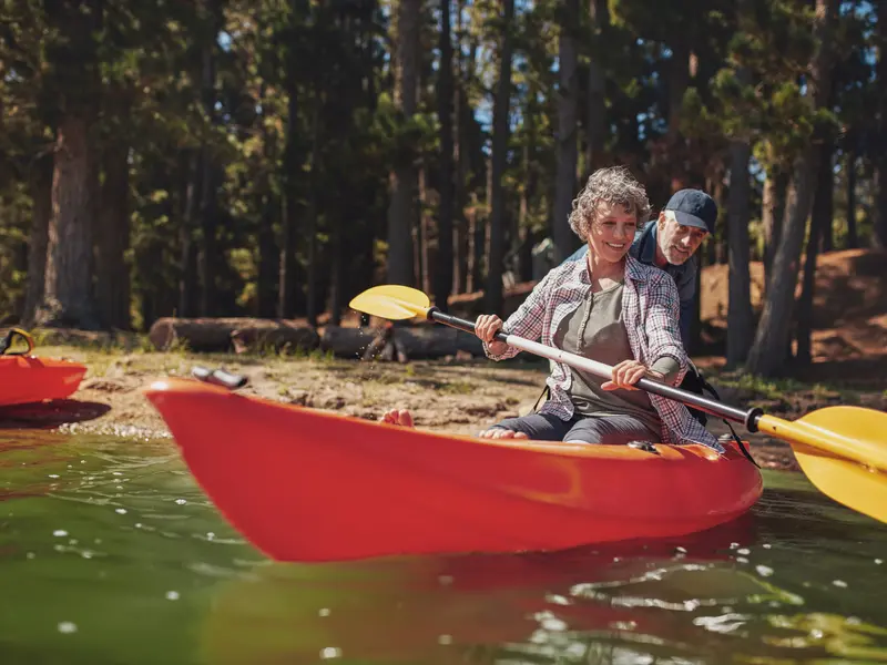 Older man with small cell lung cancer pushing a kayak off from the shore, while his wife, seated inside and holding a paddle, smiling