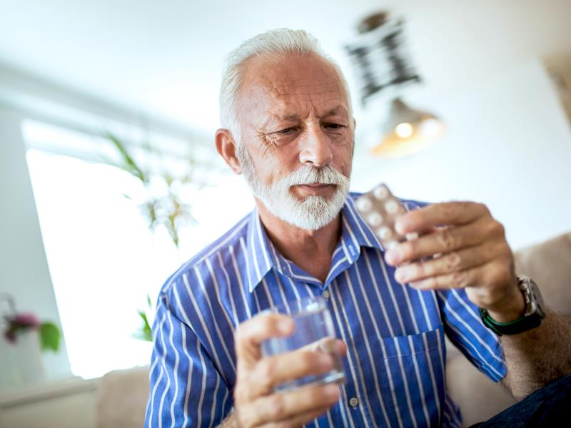 Older man at home holding a glass of water in one hand and a packet of medication in the other after an interventional cardiology procedure