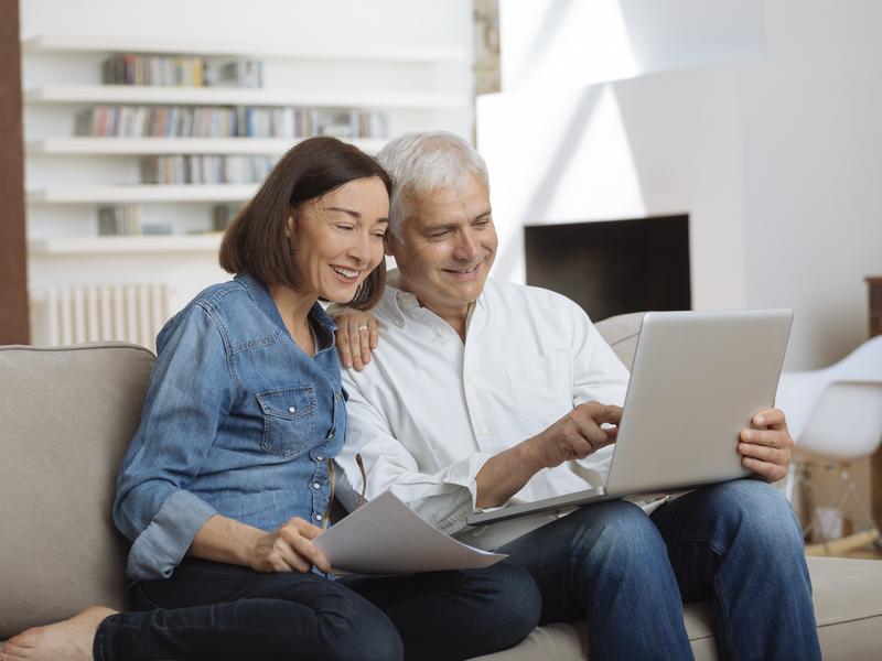 Elderly couple seated together on a couch at home, smiling and looking at a laptop while messaging their provider for health advice