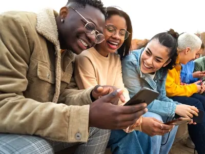 A group of three happy friends sitting outside on a phone after deciding to try a social media detox.