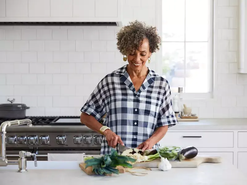 Smiling woman with curly gray hair chopping fresh vegetables on a cutting board in a bright kitchen