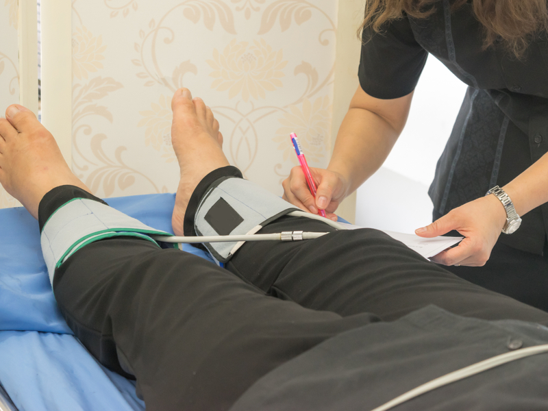 Patient lying on a medical table while a healthcare provider records the ankle-brachial index results in the patient's chart