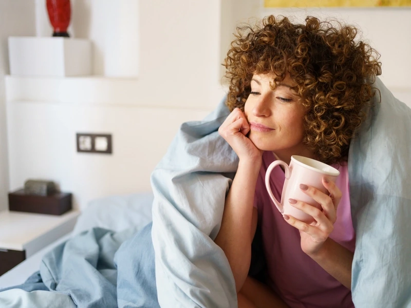 A woman in bed holding a coffee cup contemplating healthcare