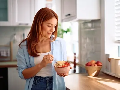 Alt tag of featured image:  A woman standing in her kitchen eating a high protein breakfast