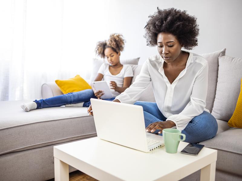 Woman sitting on a couch, opening her laptop to fill out an eVisit questionnaire while her daughter sits next to her with a tablet in hand.