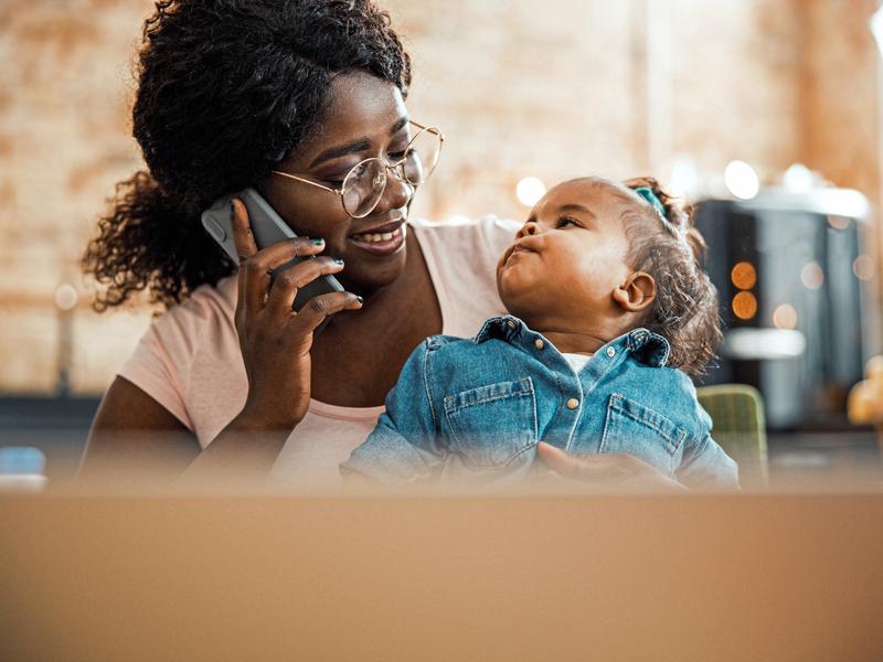 Woman smiling and conversing on her mobile phone during an online doctor visit for her two-year-old daughter, who is sitting in her lap