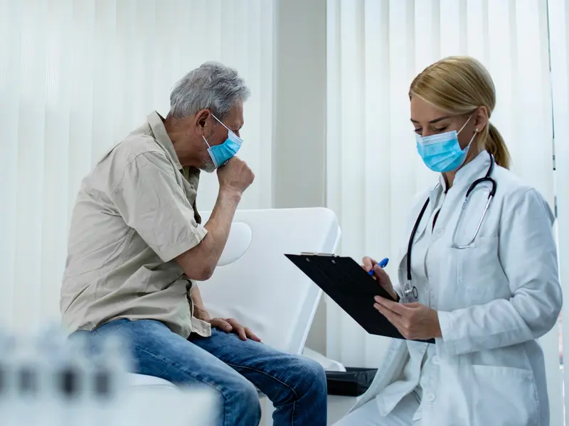 man in doctor's office with lung cancer symptoms while medical provider checks patient chart