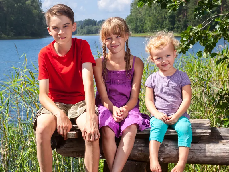 three young children sitting on wood bench in front of lake and trees smiling at camera